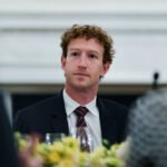 Uomo con capelli ricci e giacca scura seduto a un tavolo con decorazioni floreali.